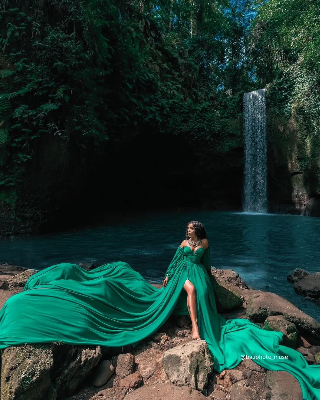 Mujer con vestido verde fluido de pie junto a una exuberante cascada en Ubud, Bali.